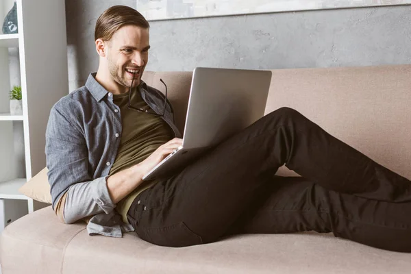 Junger glücklicher Mann mit Brille im Mund arbeitet mit Laptop auf der Couch — Stockfoto