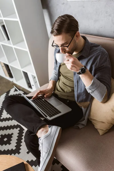 Молодой красивый мужчина работает с ноутбуком и пьет кофе на диване — стоковое фото