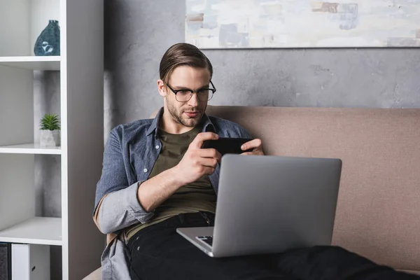 Молодой фрилансер с ноутбуком на коленях сидит на диване и играет в игру на смартфоне — стоковое фото