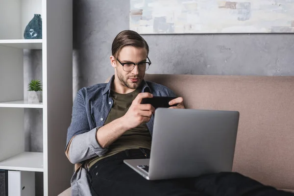 Junge Freiberuflerin mit Laptop auf Knien auf Couch sitzend und Spiel auf Smartphone — Stockfoto