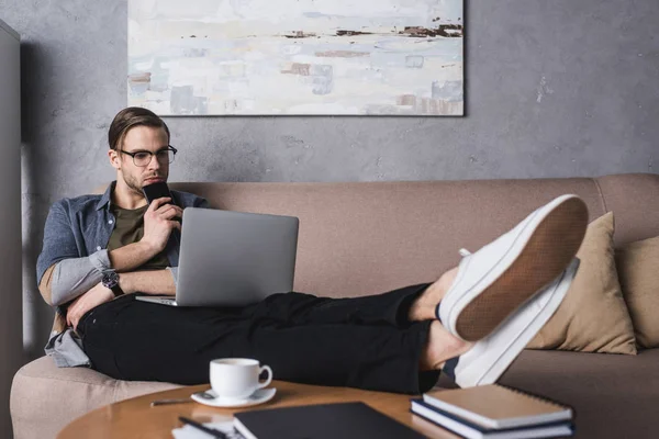 Young thoughtful man working with laptop on couch at home office — Stock Photo