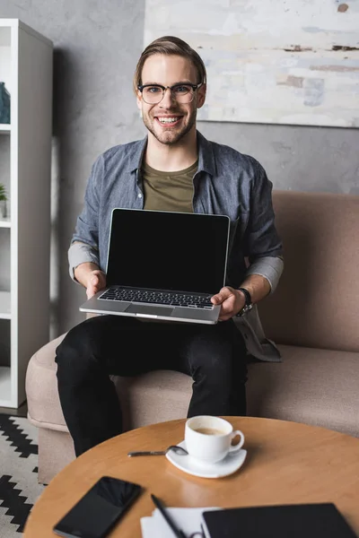 Jeune homme souriant assis sur le canapé et montrant ordinateur portable — Photo de stock