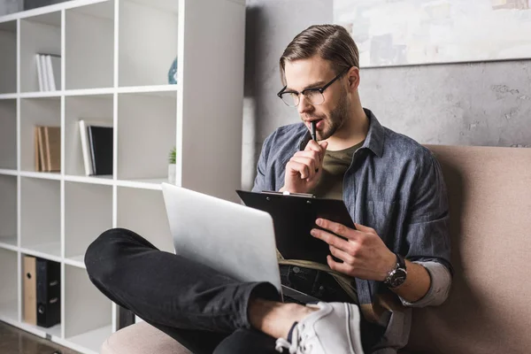 Jeune homme réfléchi qui travaille avec un ordinateur portable sur le canapé et prend des notes sur le presse-papiers — Photo de stock