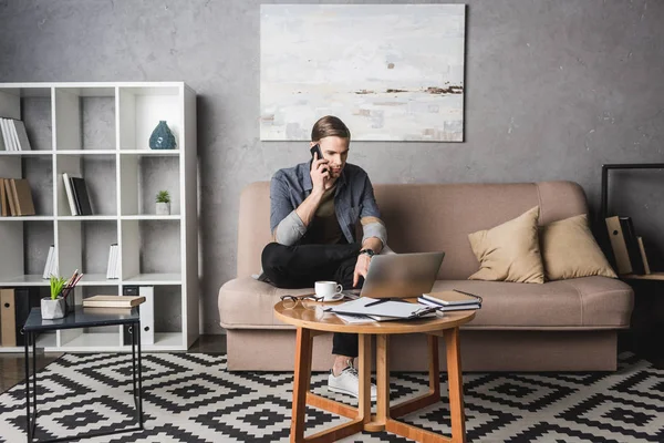 Joven hombre guapo que trabaja con el ordenador portátil en el sofá y hablando por teléfono - foto de stock