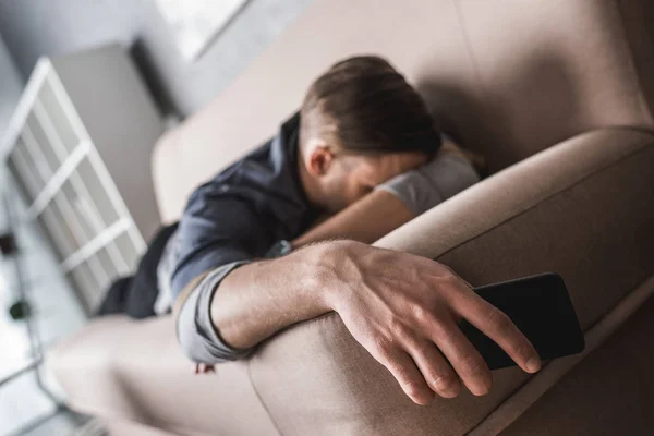 Уставший молодой человек спит на диване со смартфоном в руке — стоковое фото