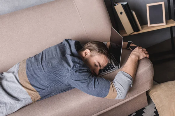 Молодой перегруженный работой бизнесмен спит на диване с головой, лежащей на ноутбуке — стоковое фото