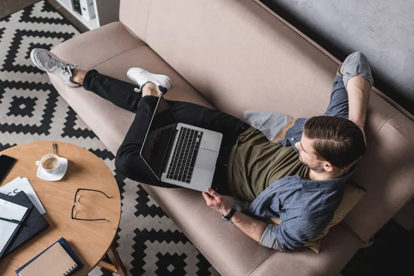 Высокий угол обзора молодого человека, работающего с ноутбуком на диване — стоковое фото