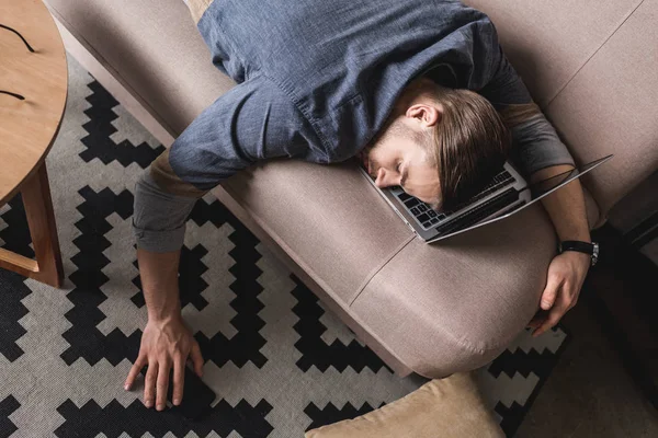 Vista de ángulo alto del hombre de negocios sobrecargado que duerme en el sofá con la cabeza acostada en el ordenador portátil - foto de stock