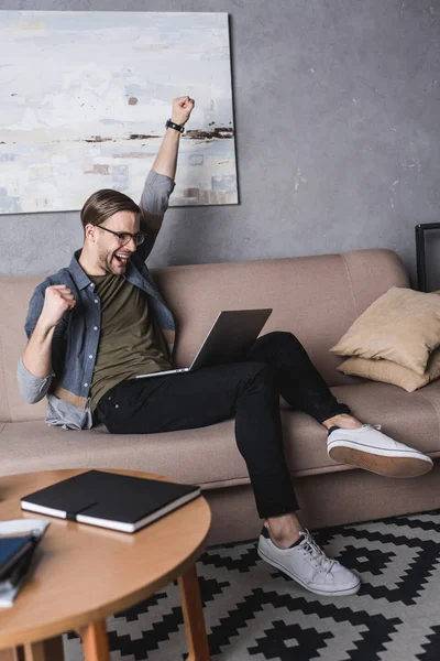 Young happy man with laptop sitting on couch and celebrating success — Stock Photo