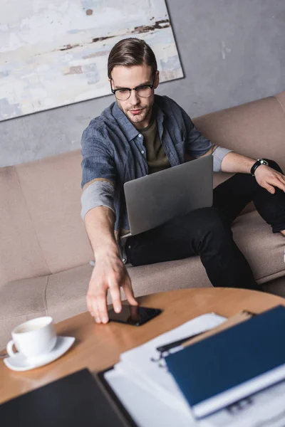 Young handsome man with laptop reaching for smartphone — Stock Photo