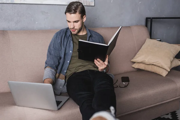 Young serious businessman reading notes on couch and using laptop — Stock Photo