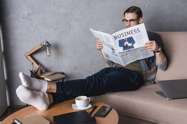 Young barefoot businessman reading newspaper on couch — Stock Photo