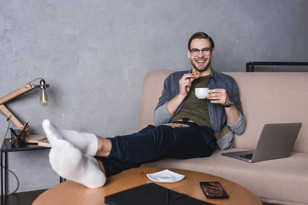 Jovem homem bonito com laptop beber café com biscoitos no sofá — Fotografia de Stock