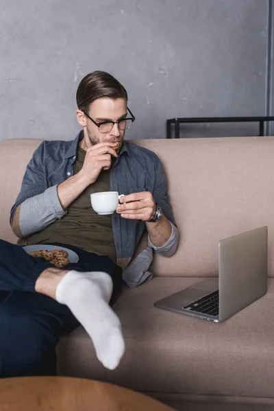 Jovem homem bonito trabalhando com laptop no sofá enquanto come biscoitos com café — Fotografia de Stock