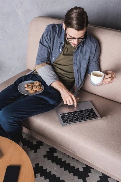 Jovem homem bonito trabalhando com laptop e beber café no sofá — Fotografia de Stock