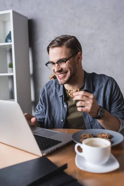 Jovem homem bonito trabalhando com laptop e comer biscoitos — Fotografia de Stock