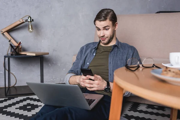 Junger skeptischer Mann blickt auf Laptop, während er auf dem Boden sitzt — Stockfoto