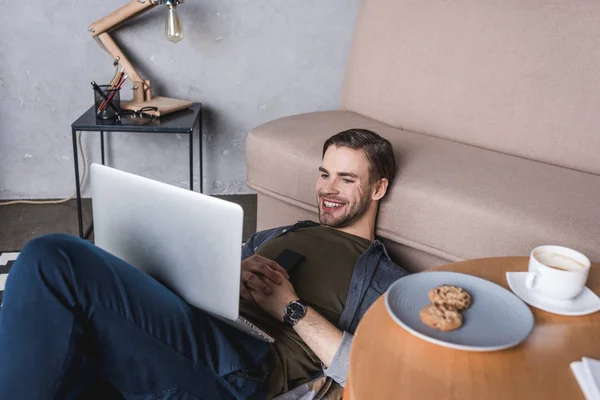 Junger glücklicher Mann benutzt Laptop, während er auf dem Boden sitzt — Stockfoto