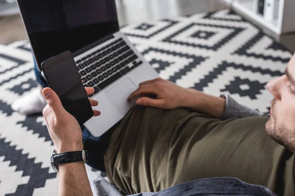 Hombre joven que trabaja con el ordenador portátil y la creación de redes con el teléfono inteligente mientras está sentado en el suelo - foto de stock