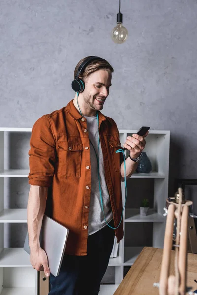 Молодой человек с ноутбуком и смартфоном слушает музыку с наушниками — стоковое фото