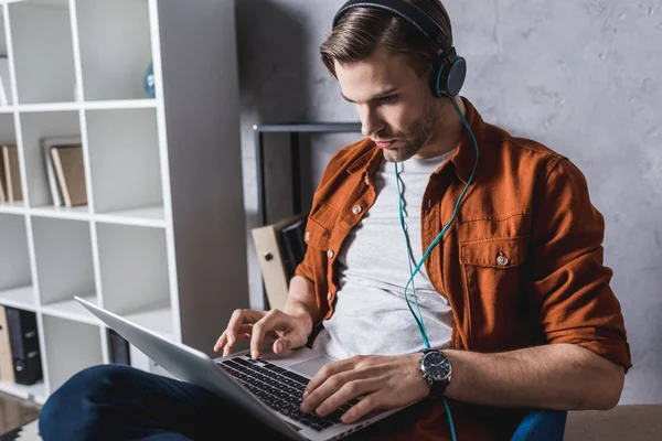 Joven hombre guapo en los auriculares que trabajan con el ordenador portátil en el sofá - foto de stock