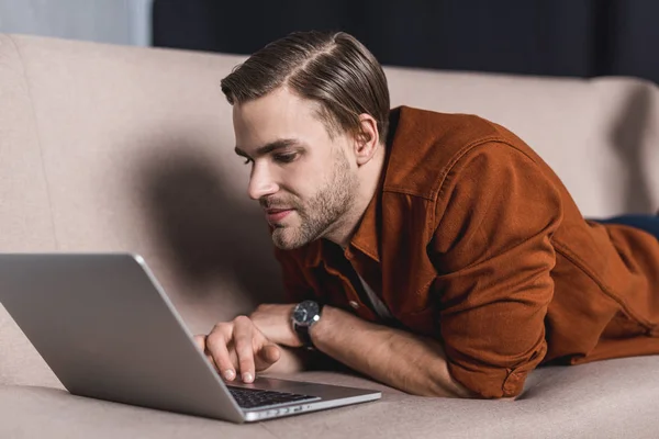 Молодой улыбающийся мужчина работает с ноутбуком на диване — стоковое фото