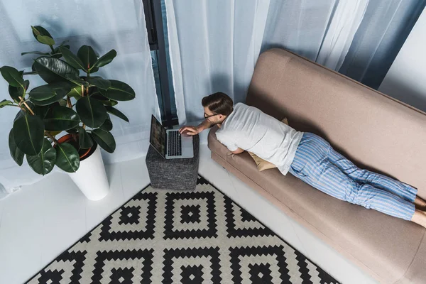 Visão de alto ângulo de freelancer em pijama trabalhando com laptop no sofá — Fotografia de Stock