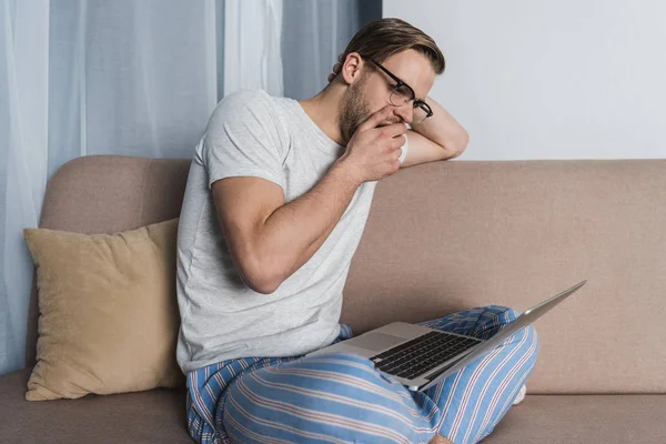 Зевающий перегруженный фрилансер в пижаме, работающий с ноутбуком на диване — стоковое фото
