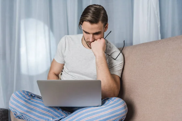 Joven freelancer reflexivo en pijama trabajando con el ordenador portátil en el sofá - foto de stock