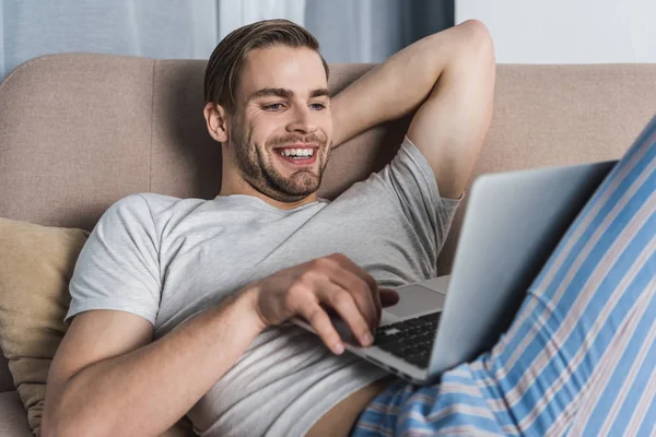 Jeune freelance souriant en pyjama travaillant avec ordinateur portable sur le canapé — Photo de stock