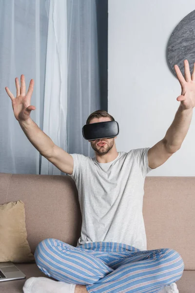 Joven en pijama y casco de realidad virtual sentado en el sofá - foto de stock