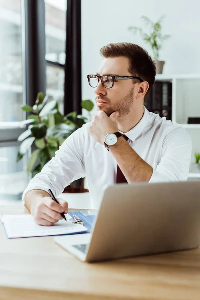 Handsome pensive businessman with paperwork and laptop at workspace — Stock Photo