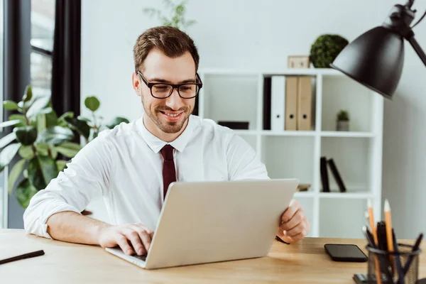 Apuesto hombre de negocios sonriente trabajando con el ordenador portátil en la mesa con el teléfono inteligente - foto de stock