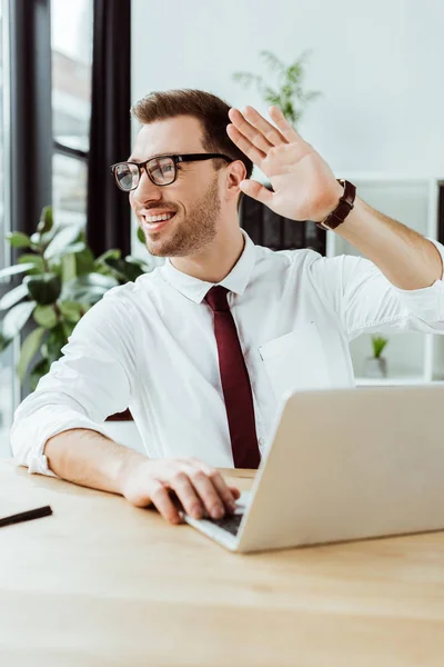 Hombre de negocios sonriente trabajando con el ordenador portátil en el lugar de trabajo y saludando a alguien — Stock Photo