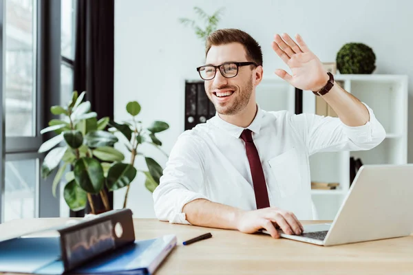 Cheerful businessman waving and using laptop at workplace — Stock Photo