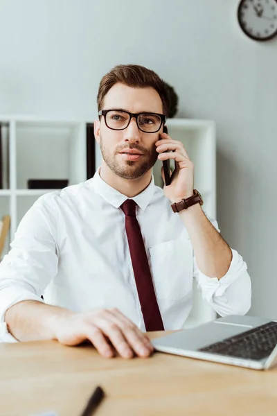 Hombre de negocios guapo hablando en el teléfono inteligente mientras está sentado en la oficina con el ordenador portátil - foto de stock
