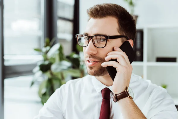 Schöner Geschäftsmann mit Brille, der im Büro auf dem Smartphone spricht — Stockfoto