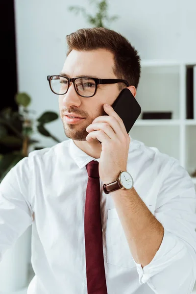 Guapo hombre de negocios hablando en el teléfono inteligente mientras trabaja en la oficina - foto de stock