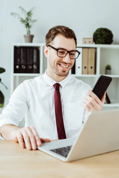 Handsome businessman using smartphone and laptop at workplace in modern office — Stock Photo