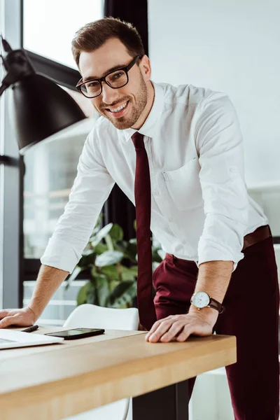 Apuesto ejecutivo alegre hombre de negocios de pie en la mesa con el teléfono inteligente - foto de stock