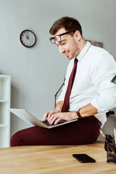 Cheerful businessman using laptop while sitting on table in office — Stock Photo