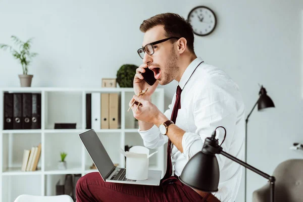 Businessman working with smartphone and laptop while eating takeout noodles for lunch in office — Stock Photo