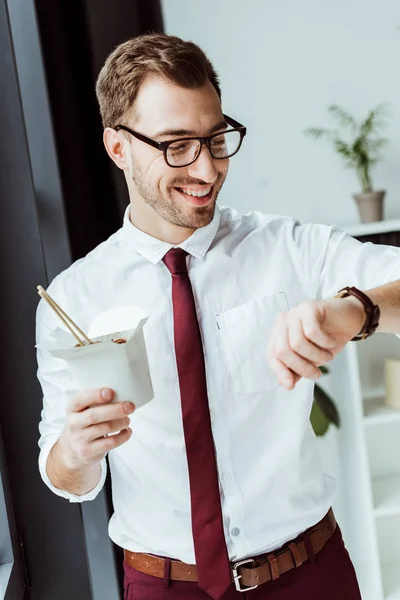 Sonriente hombre de negocios sosteniendo caja con fideos y mirando el reloj — Stock Photo