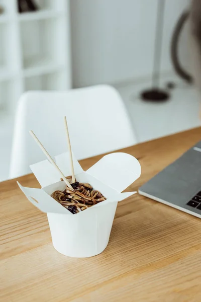Коробка для еды с лапшой и палочками на рабочем месте с ноутбуком — стоковое фото