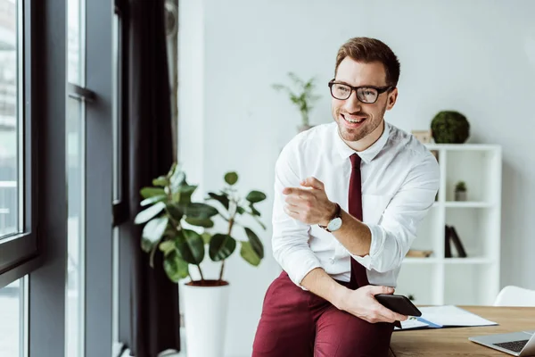 Schöner Geschäftsmann mit Brille, Smartphone in der Hand und auf jemanden zeigend — Stockfoto