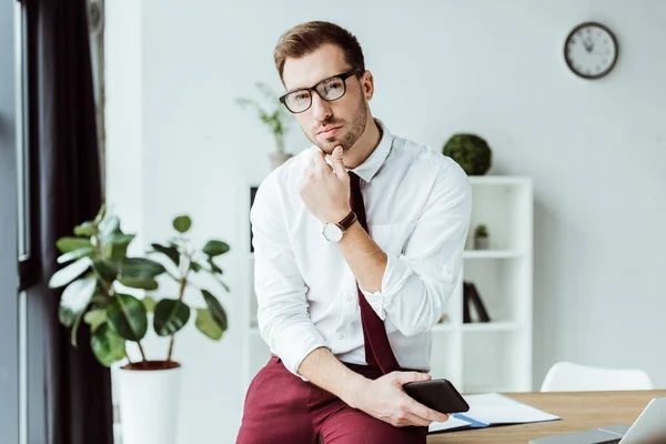 Handsome pensive businessman in eyeglasses holding smartphone at workplace — Stock Photo