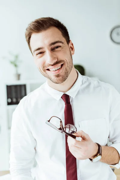Cheerful handsome businessman holding eyeglasses and looking at camera — Stock Photo