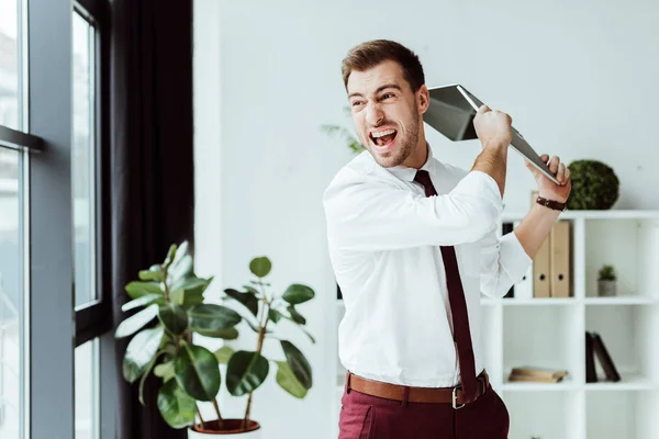 Aggressive businessman screaming and throwing laptop in modern office — Stock Photo