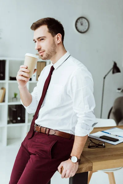 Pensativo hombre de negocios con café para ir apoyándose en la mesa en la oficina moderna - foto de stock