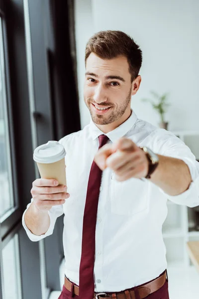 Hombre de negocios sonriente con taza de café desechable apuntando a usted — Stock Photo